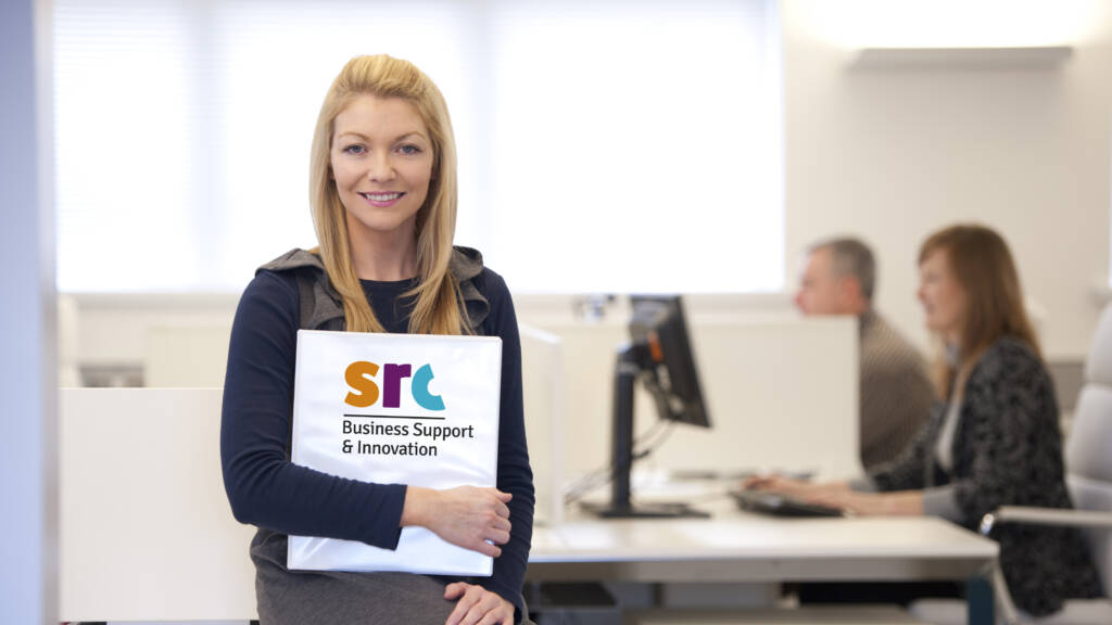 A female members of the Business Support Team is sitting on a desk within an office, she is smiling and holding a document labelled SRC Business Support and Innovation. Two other members of the team are working on desktop computers in the background.