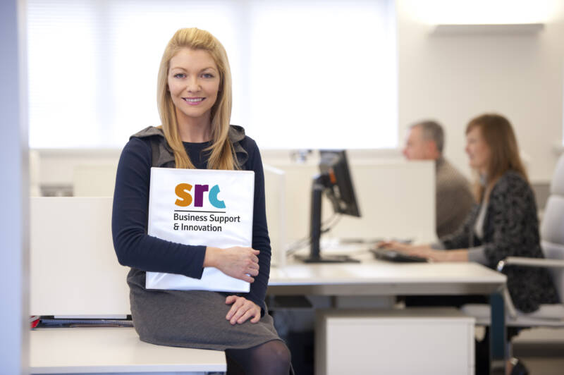 A female members of the Business Support Team is sitting on a desk within an office, she is smiling and holding a document labelled SRC Business Support and Innovation. Two other members of the team are working on desktop computers in the background.
