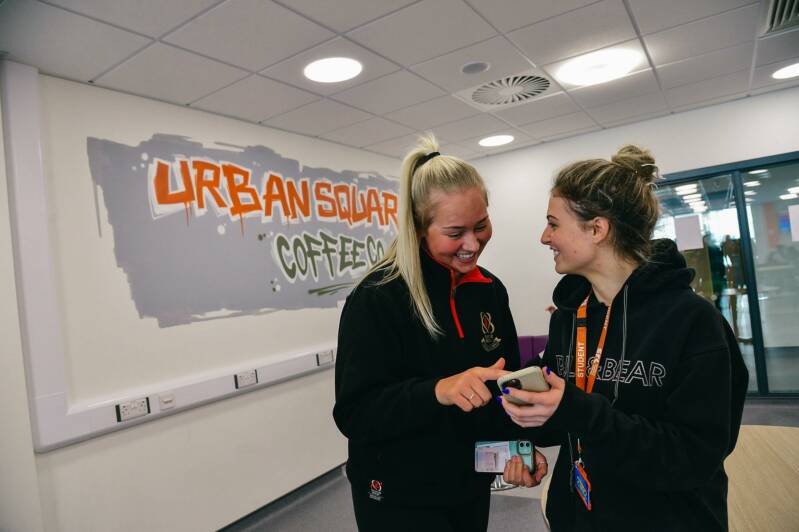 Two female students are standing and chatting in front of SRC Urban Square Coffee Co signage on wall