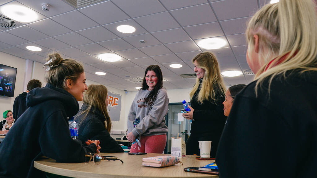 A group of SRC students are smiling and chatting round a table in a relaxed casual seating area. There are coffee cups and mobile phones on the table and in their hands.