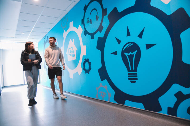 Two students walking along corridor, the wall is bright blue with large decals of lightbulbs, cogs and other tech related symbols