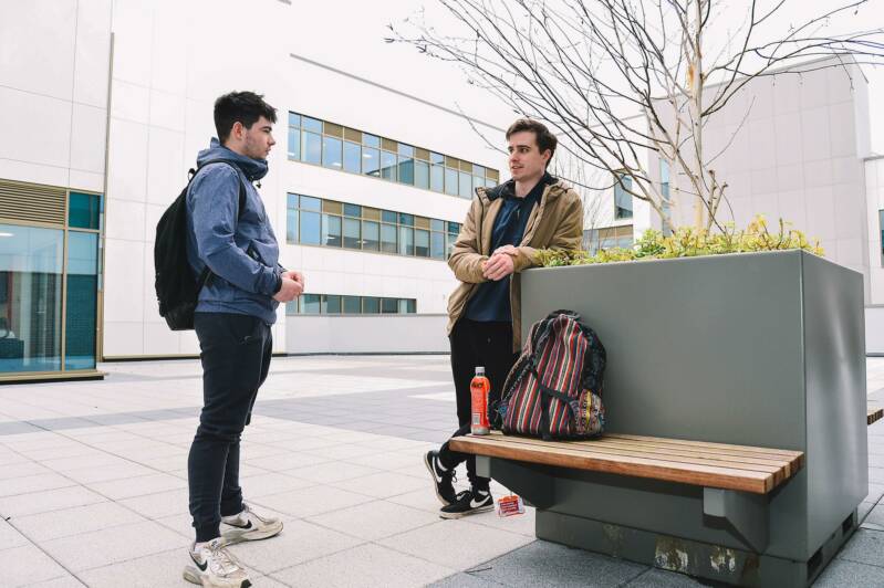 Two male students are relaxing outside the SRC Armagh Campus, one is leaning on a large planter which has a bench attached.
