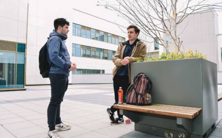 Two male students are relaxing outside the SRC Armagh Campus, one is leaning on a large planter which has a bench attached.