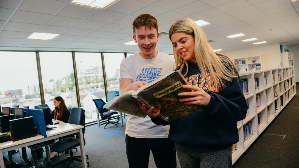 A male and female student are sharing a book and looking at a page together in an SRC learning resource area. There are rows of bookshelves in the background and another student is working on a desktop computer.