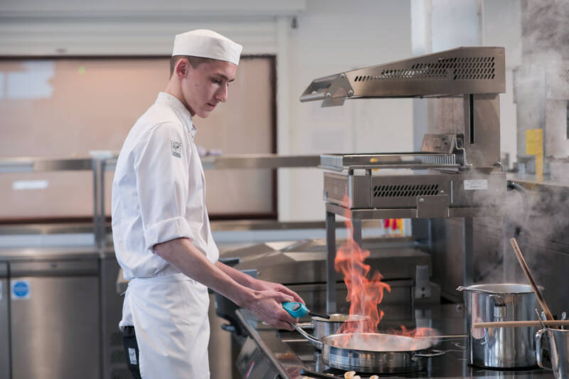 Trainee chef working in restaurant kitchen with flaming saucepan