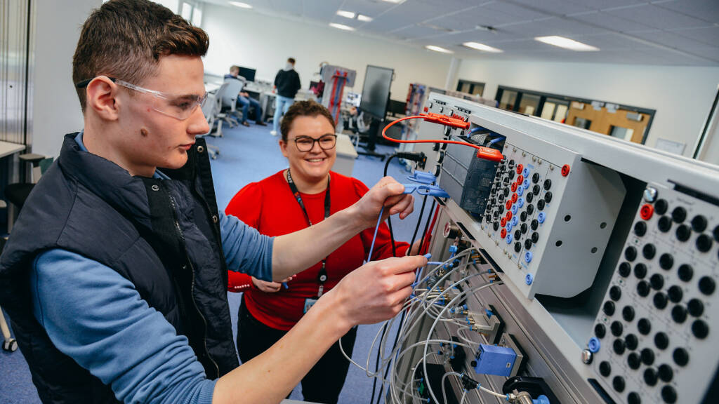 An SRC student is working with a large electronic system in an electronic engineering lab while a member of SRC staff looks on, smiling. They are both wearing safety glasses.