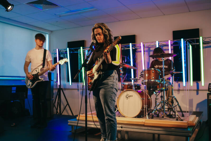 Band playing in SRC music studio including a drummer on a drumkit, a student playing an electric guitar and another student playing an electric guitar and singing into a microphone.