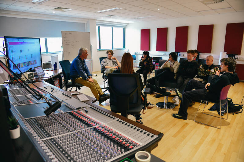 Group of SRC students and staff member sitting on chairs in a circle in music studio during class session. There is a large mixing desk in the foreground and a large digital screen at the back of the room.