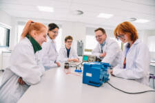 SRC science students wearing lab coats are gathered around a bench within a science lab