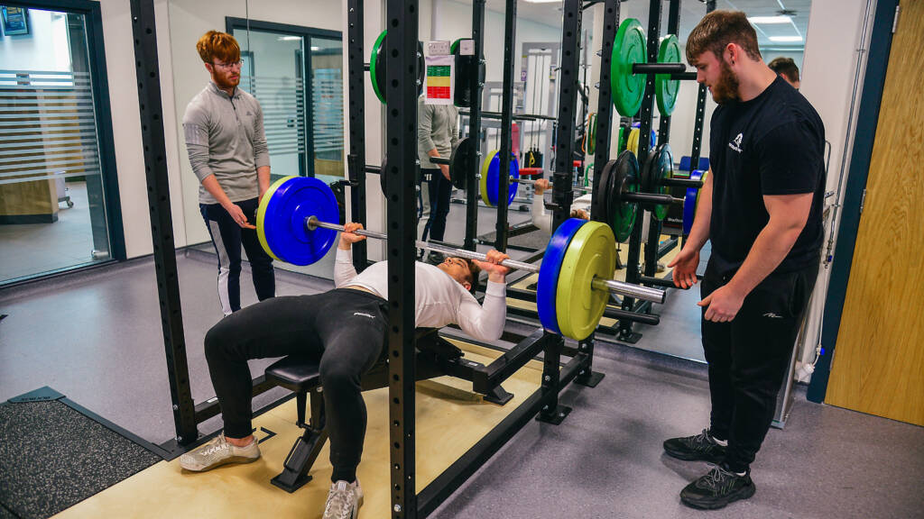 A male SRC student is completing bench presses on a weights bench while two other students assist.