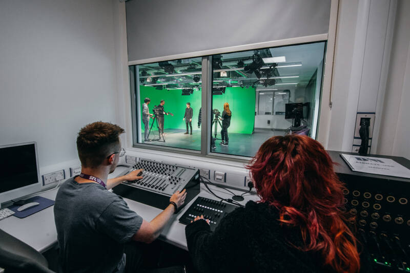 Group of students using media studio with mixing desk, AV equipment and green screens.